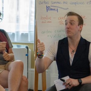 A teacher squats in front of participants with eager faces. Behind them, several signs with Latin phrases defined in Latin cover the walls.