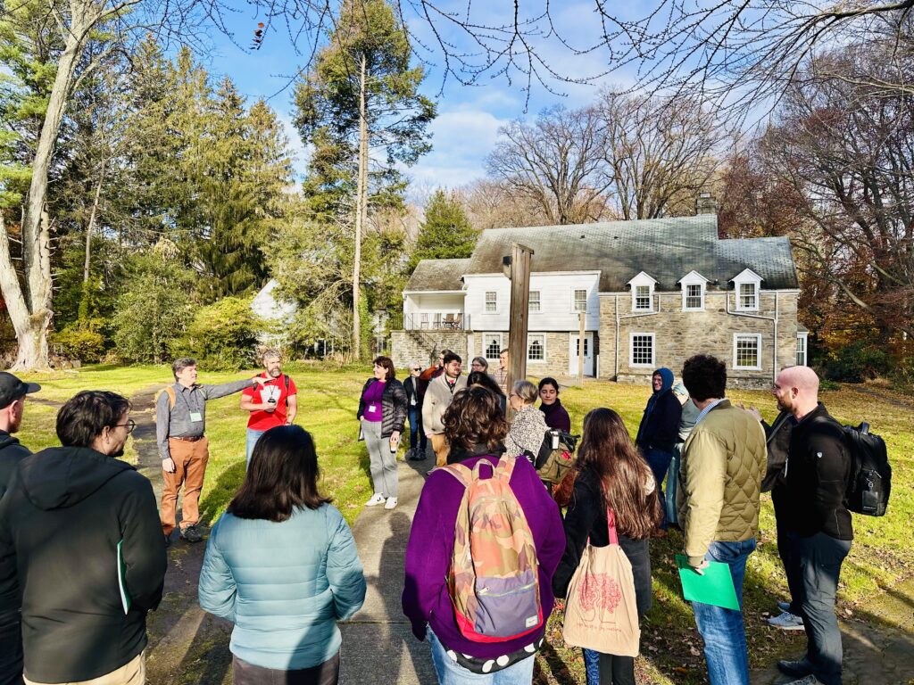A circle of people stand outside in front of a historic building and cluster of trees, listening to a man speak.