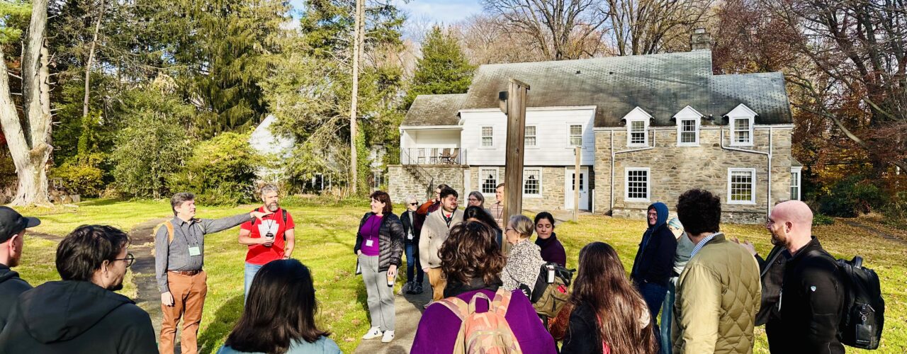 A circle of people stand outside in front of a historic building and cluster of trees, listening to a man speak. 