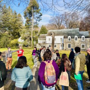 A circle of people stand outside in front of a historic building and cluster of trees, listening to a man speak.